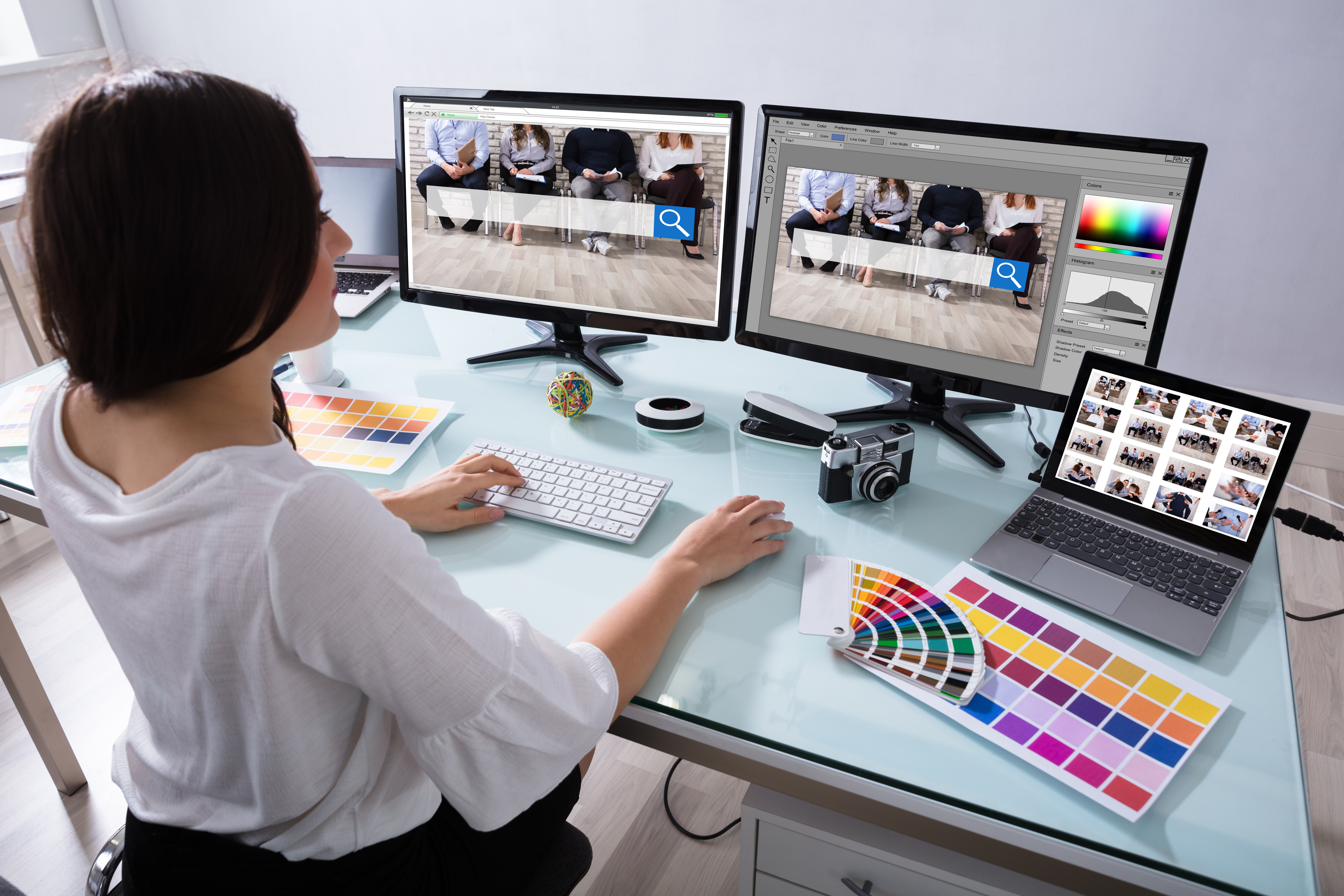 Female designer in home office sitting at her desk and looking at two monitors that show a final website image and one in a photo editing software.