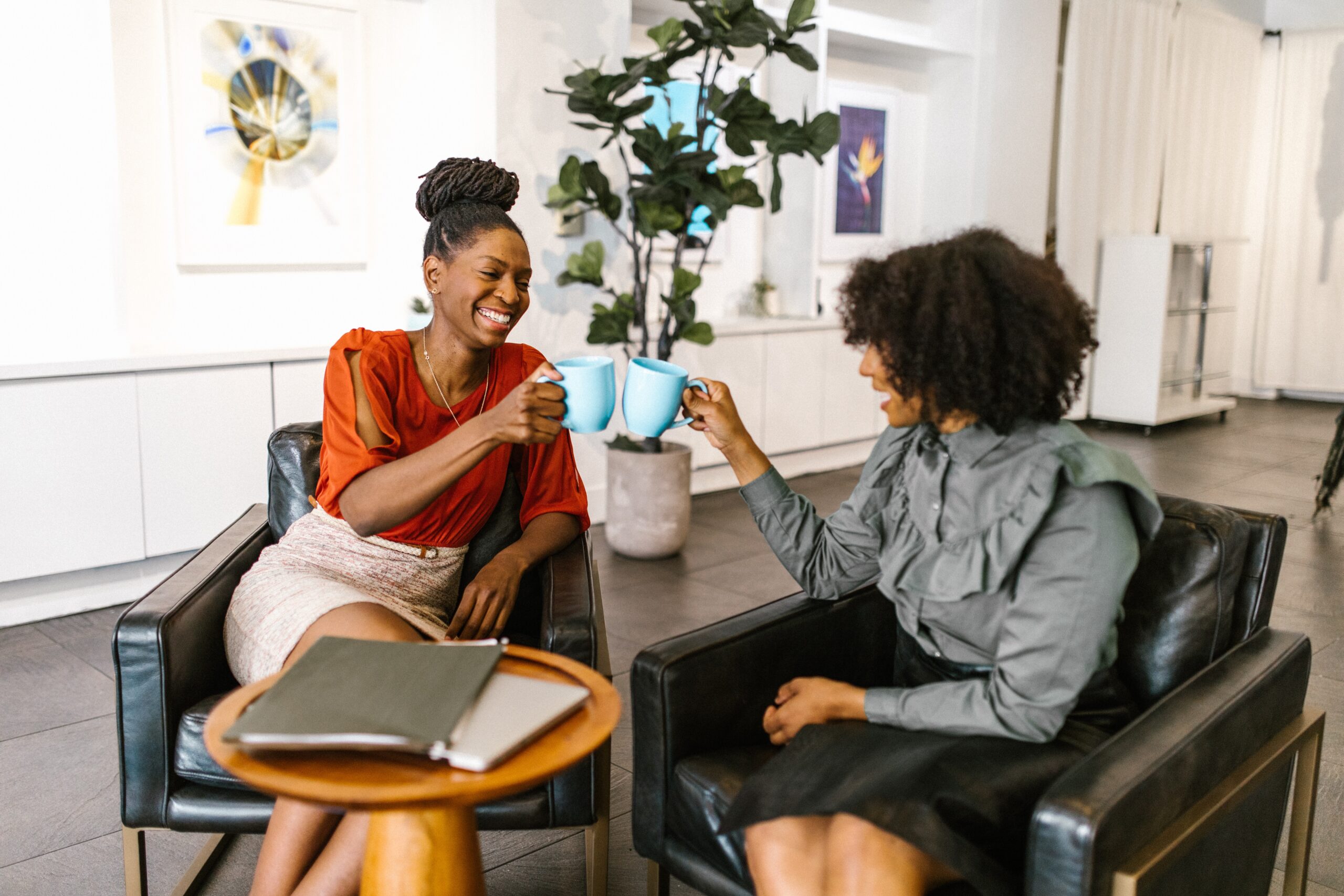 Two smiling women sitting on sofa chairs and toasting their blue coffee mugs.